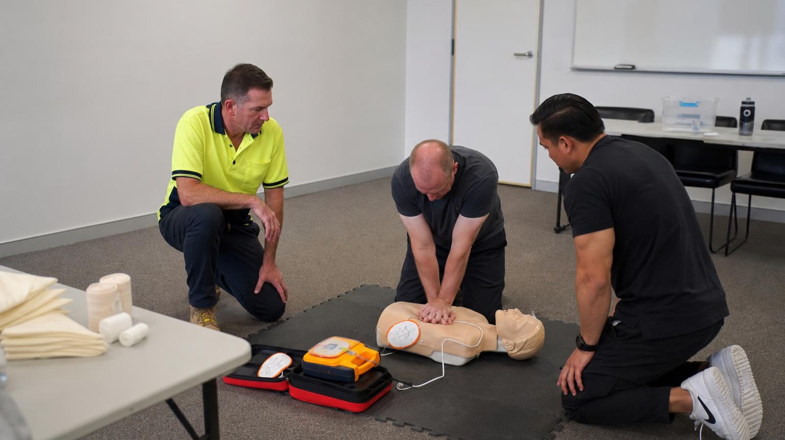 First aid course Perth candidate performing HLTAID011 CPR compressions on an adult manikin with AED trainer beside, assessor observing in hi-vis polo